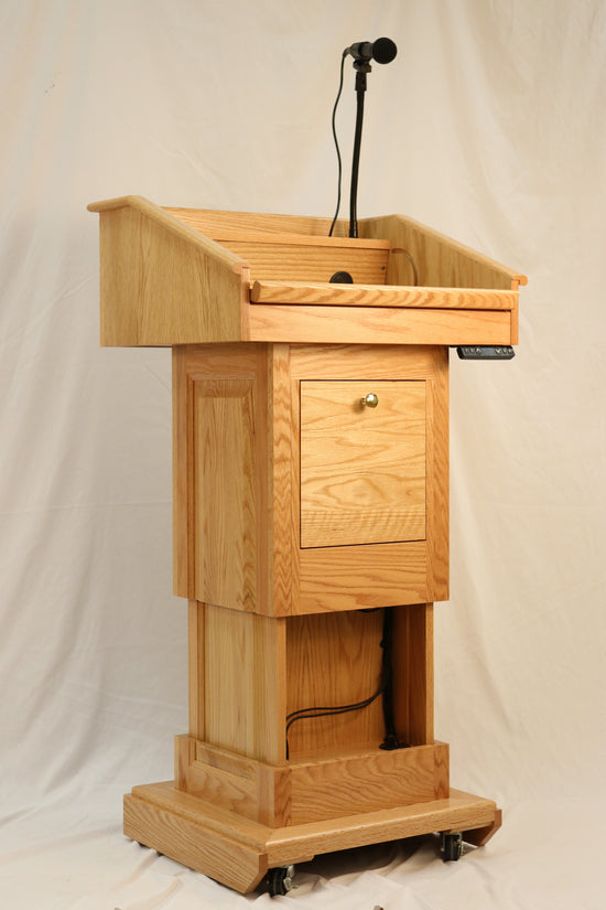 The Counselor Evolution Lift™ lectern, handcrafted from oak wood, features adjustable height, built-in sound system, and mobility casters.