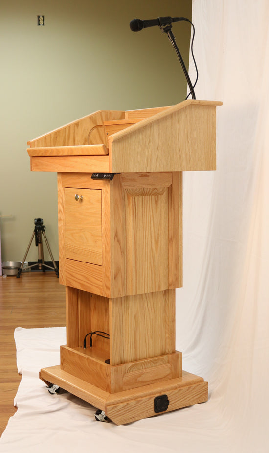 The Counselor Evolution Lift™ lectern, handcrafted from oak wood, features adjustable height, built-in sound system, and mobility casters.